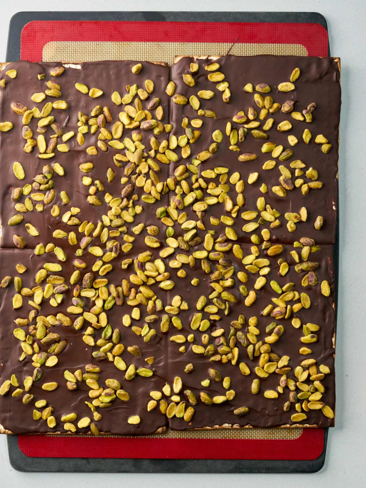 A large square matzo covered in chocolate and pistachios on top of a baking mat.