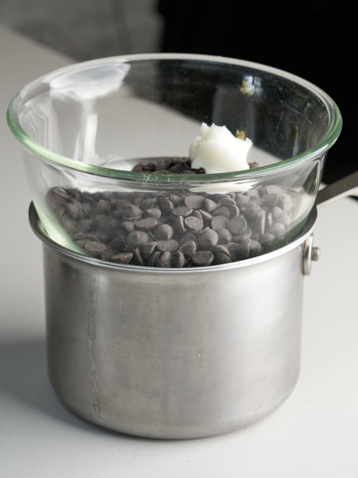 A glass bowl of chocolate chips and a dollop of coconut oil resting on a large silver saucepan.