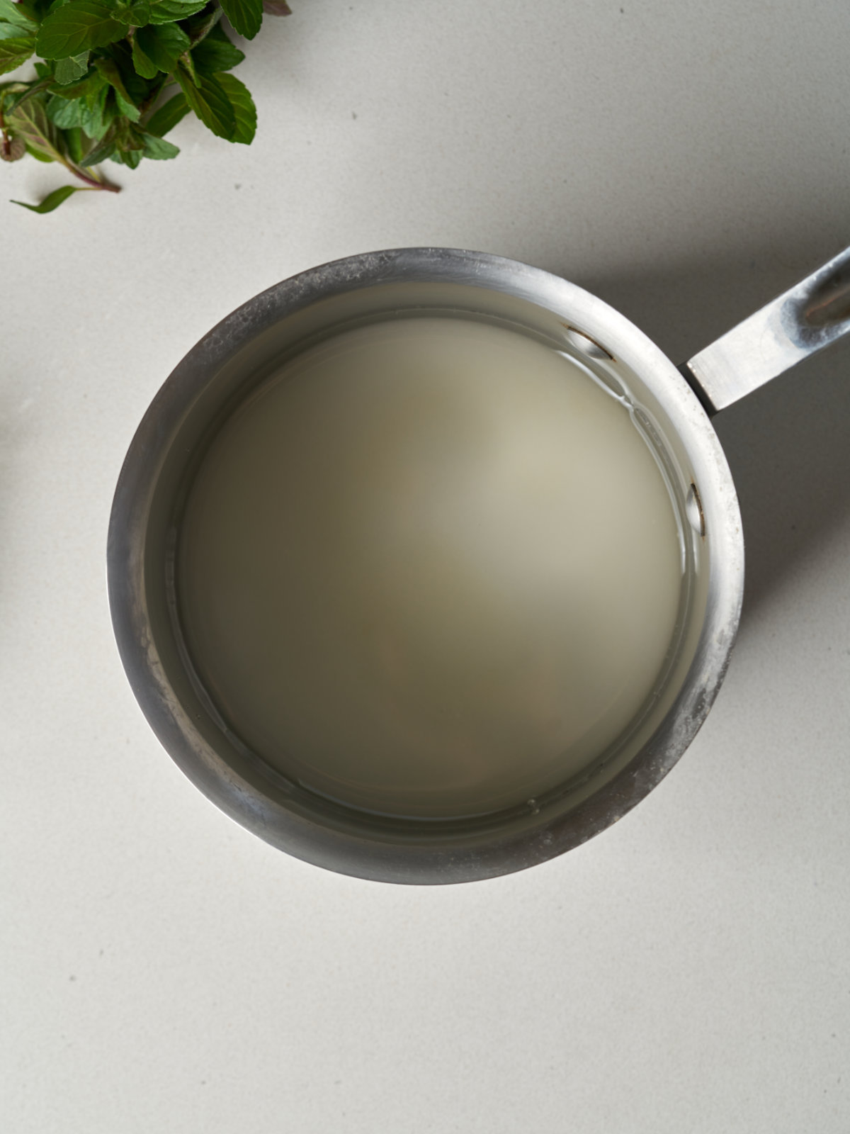 Sugar and water in a saucepan next to some mint leaves.