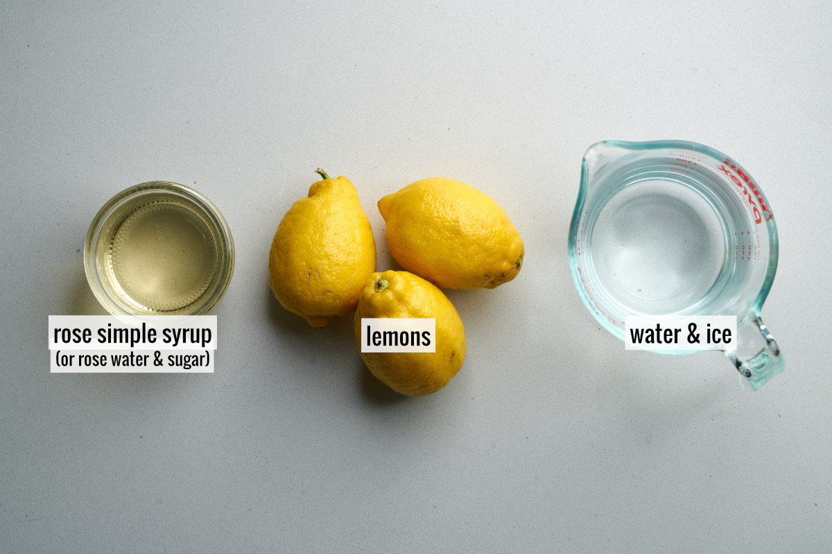 Three lemons next to a liquid measuring cup with water and a small jar of yellow tinted simple syrup.