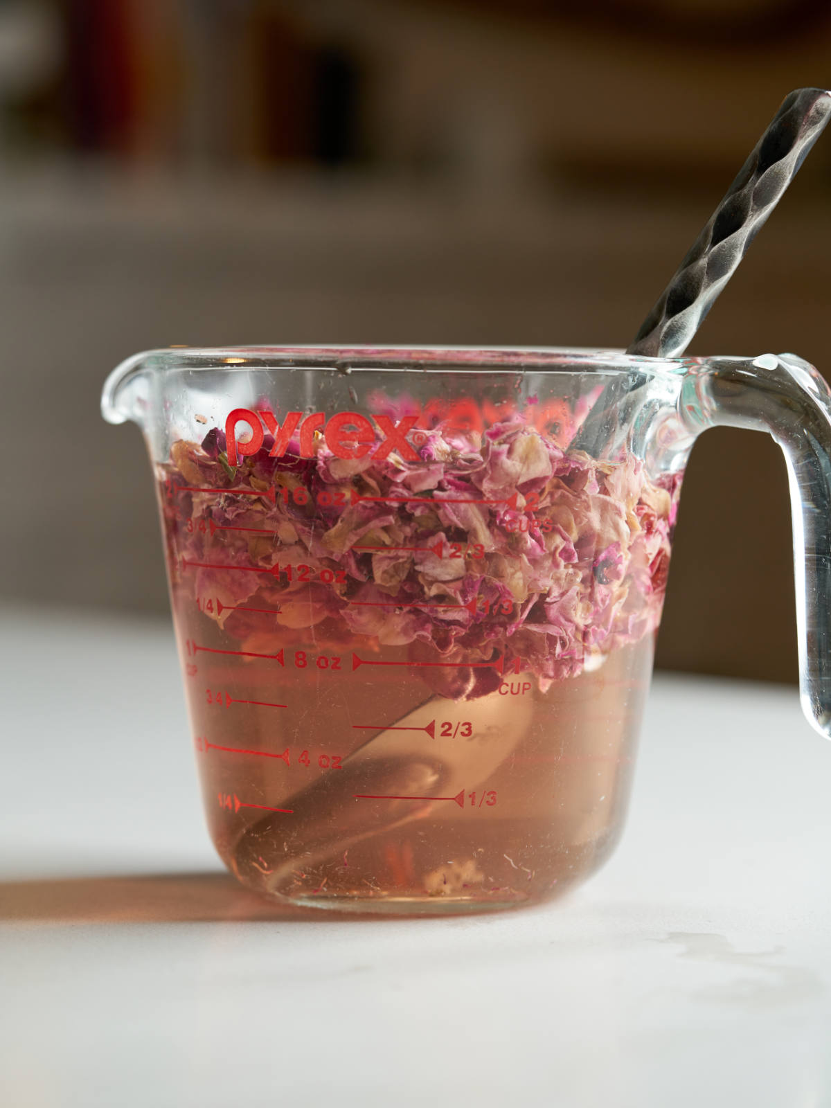 Rose petals steeped in tinted water in a liquid measuring cup with a metal spoon.