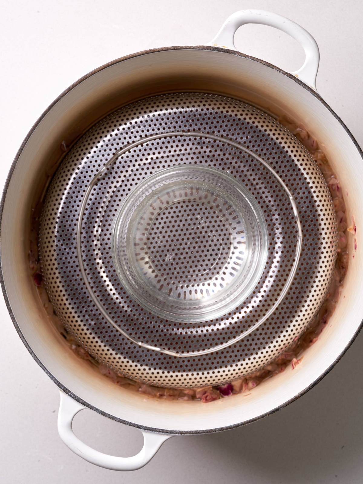 White pot filled with an inverted metal strainer and topped with a small glass bowl with cooked tiny rose petals surrounding the metal strainer.