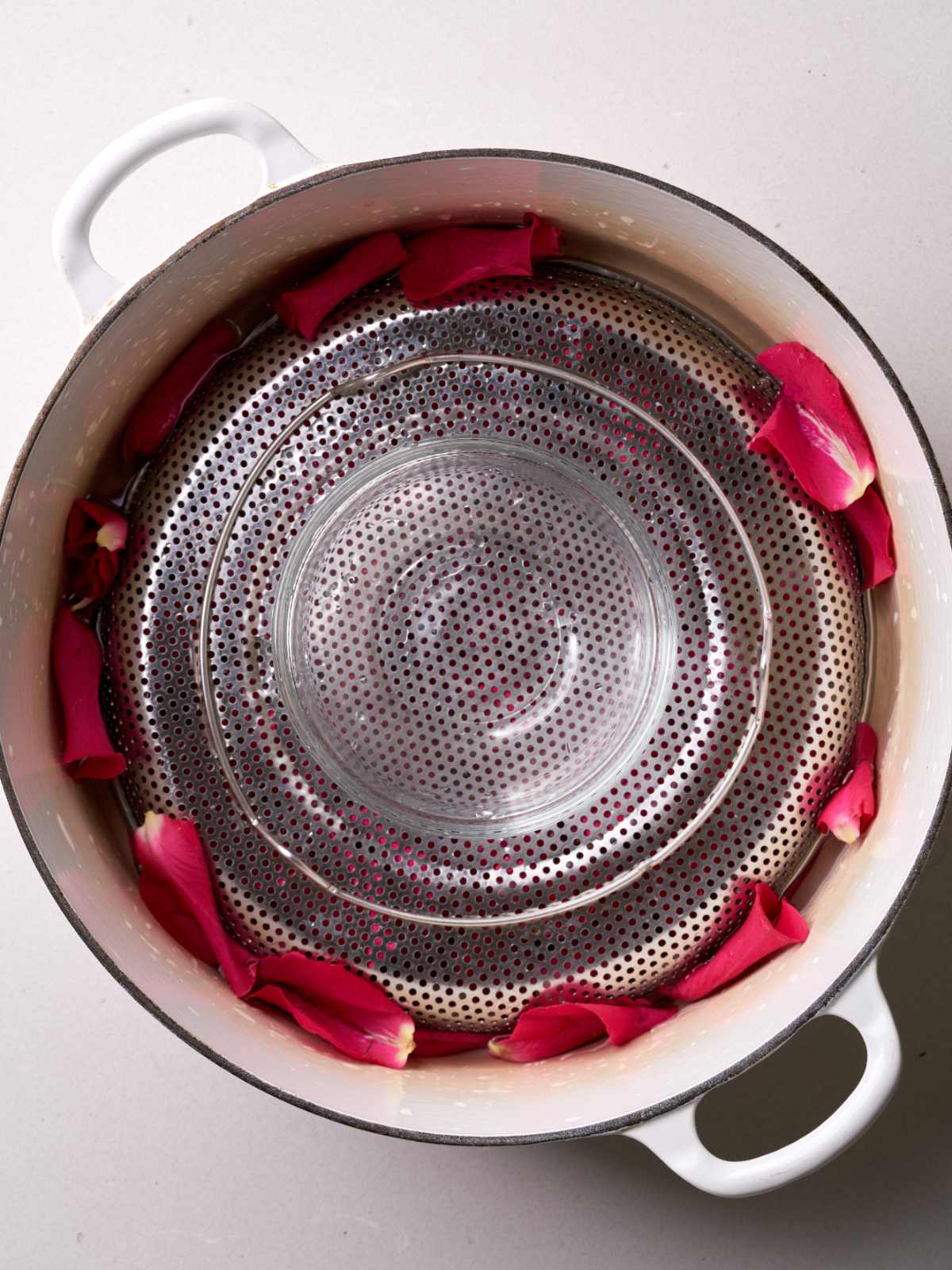 White pot filled with an inverted metal strainer surrounded by rose petals and topped with a glass bowl.