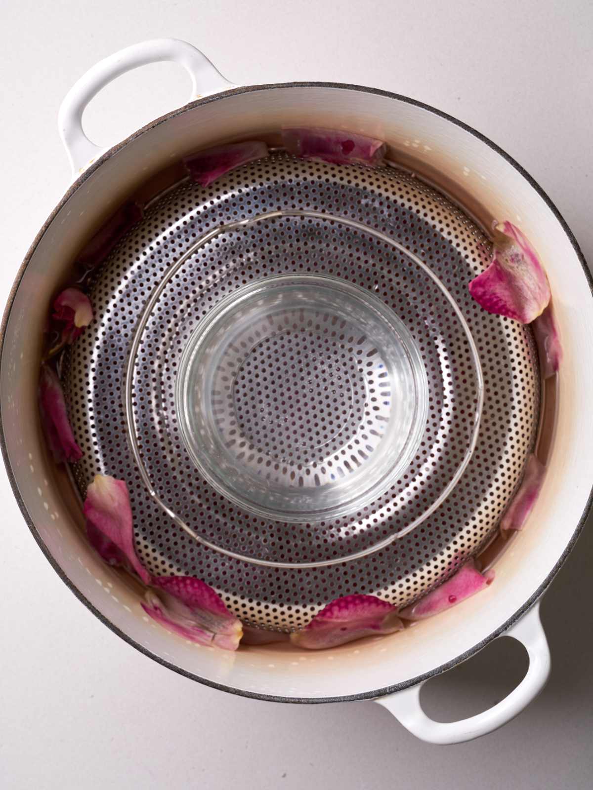 White pot with cooked rose petals topped with a metal strainer and a glass bowl with rose water.