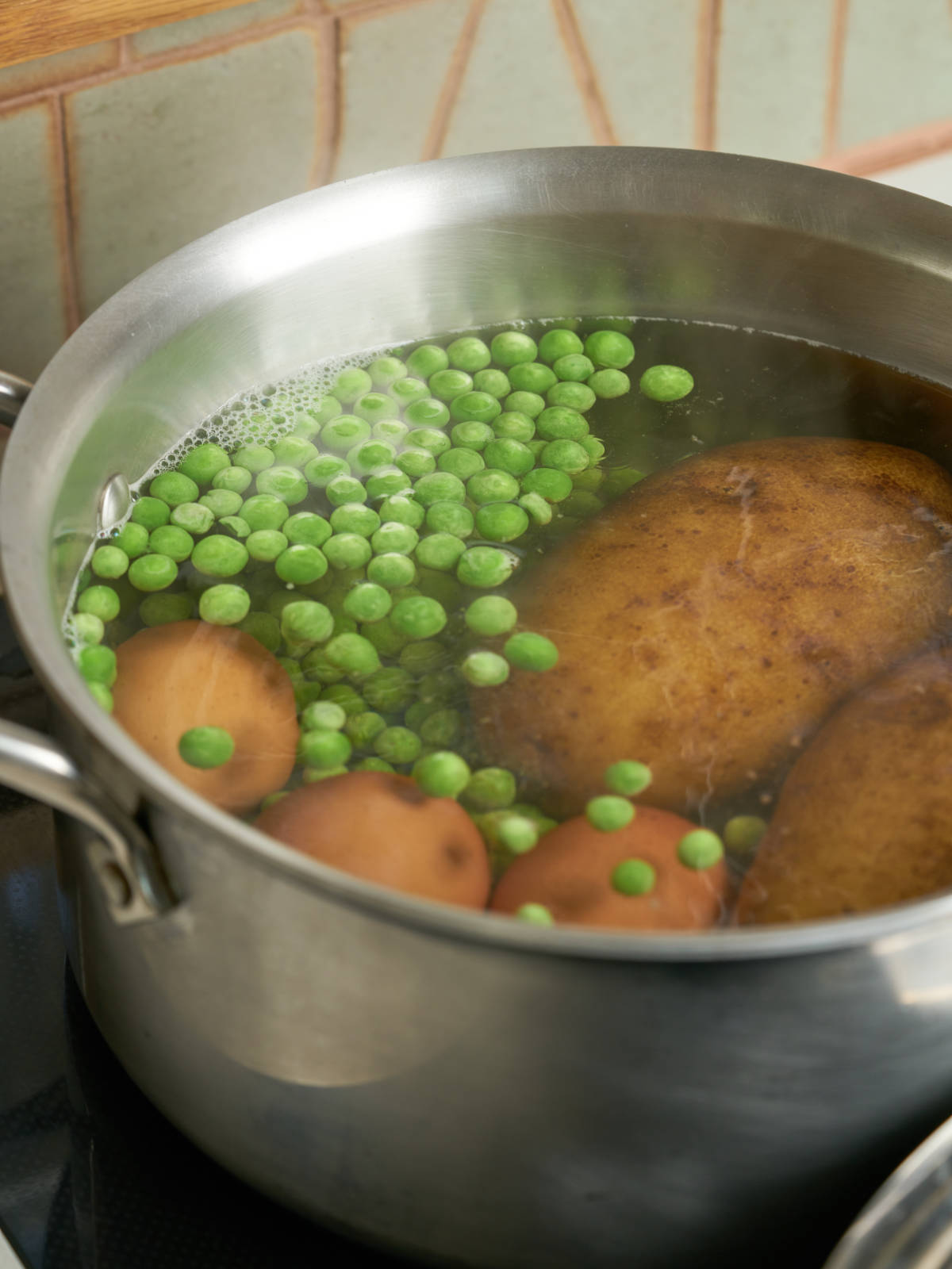 Eggs, green peas, and potatoes submerged in water in a large silver saucepan.