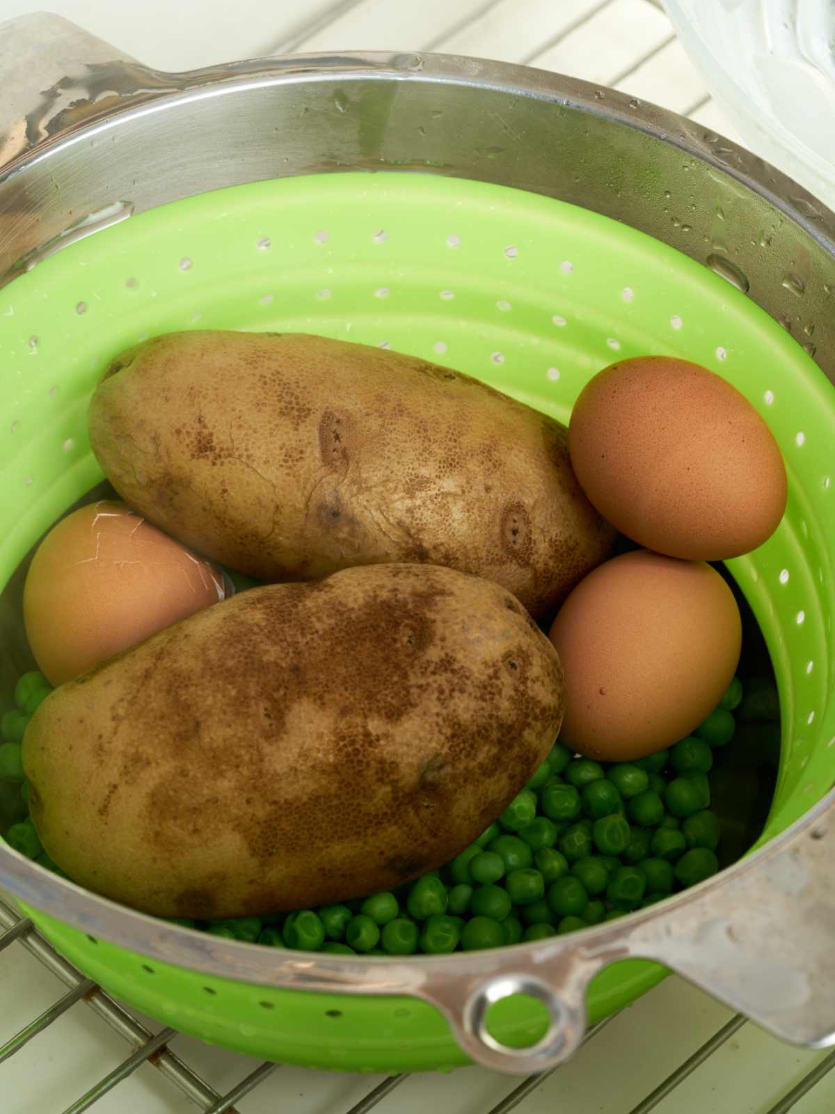 Potatoes, eggs, and green pees straining in a green colander over a silver pan.