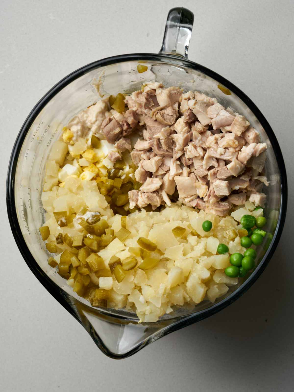 Diced chicken, potatoes, peas, and pickles in a large glass measuring jug.