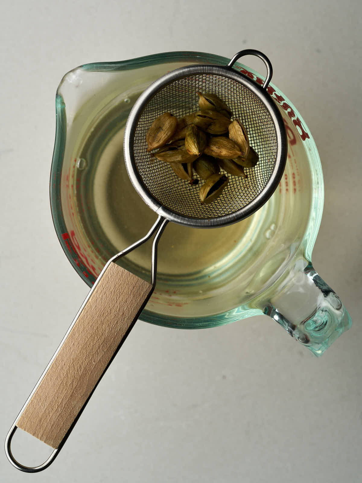 Strainer with cardamom pods over a light yellow liquid in a measuring cup.