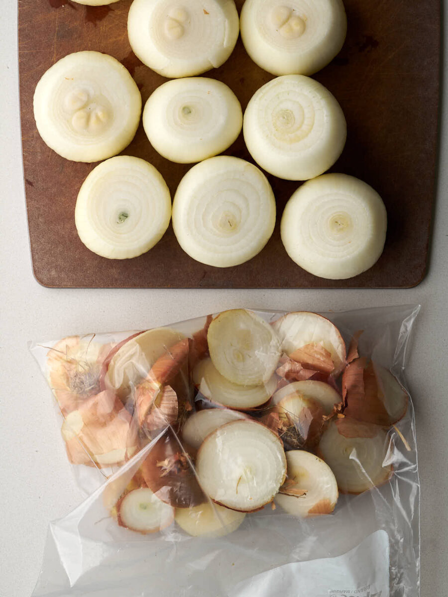 Raw onions on a wood cutting board with the discarded skins in a ziplock bag.