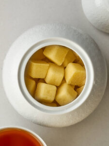 Yellow sugar cubes in a sugar bowl next to the lid and a cup of tea.
