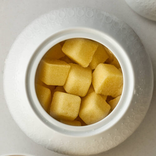 Yellow sugar cubes in a sugar bowl next to the lid and a cup of tea.