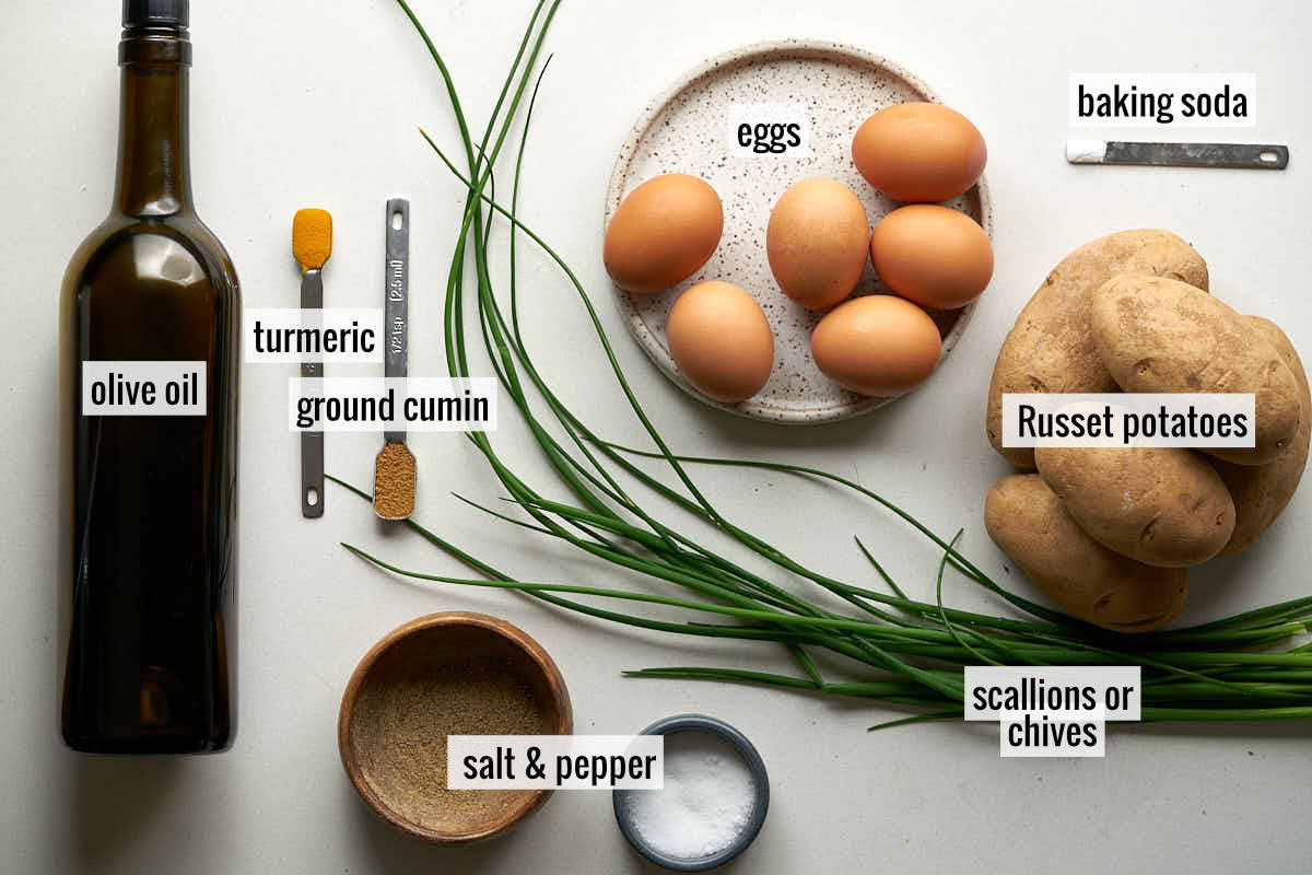 Ingredients laid out on a white surface: a dark bottle of oil, measuring spoons with turmeric and another spice, a bundle of chives, a plate of eggs, a small dish of salt, a wooden bowl with another spice, and a pile of potatoes.