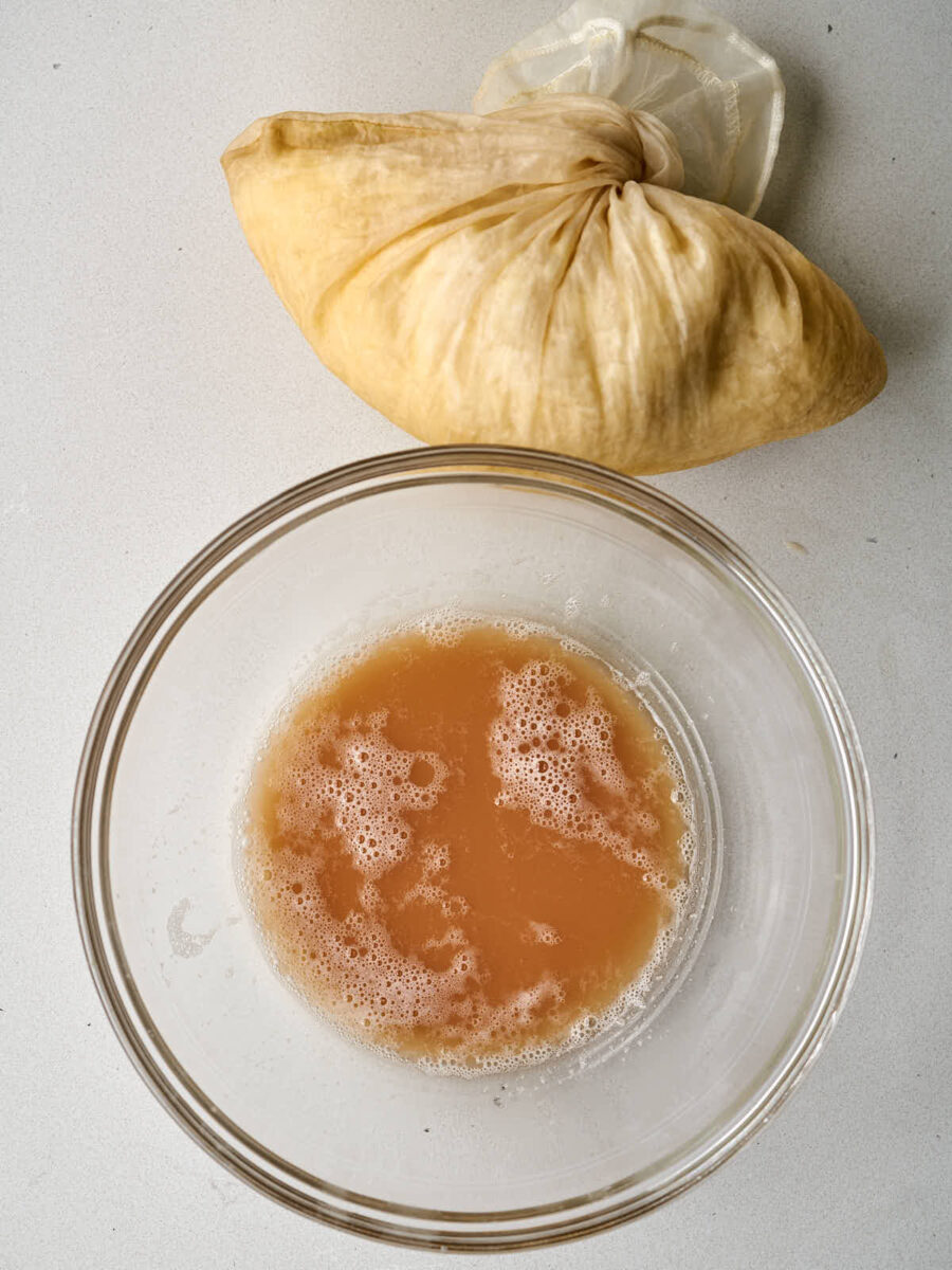 A clear glass bowl containing brown liquid next to a filled nut milk bag.