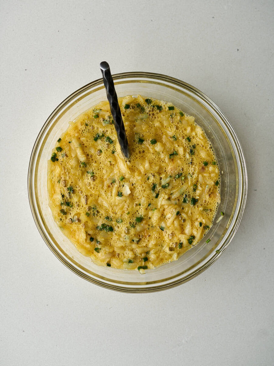 A clear glass bowl containing a mixture of beaten eggs, shredded potatoes, and chopped green onions. A black spoon is stuck into the mixture.