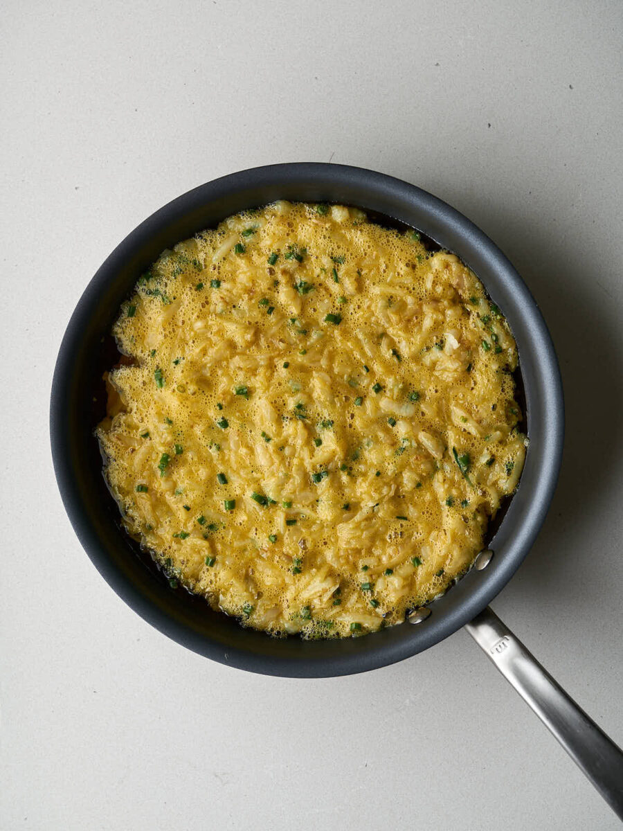 A black frying pan filled with the mixture of beaten eggs, shredded potatoes, and chopped green onions, ready to be cooked.