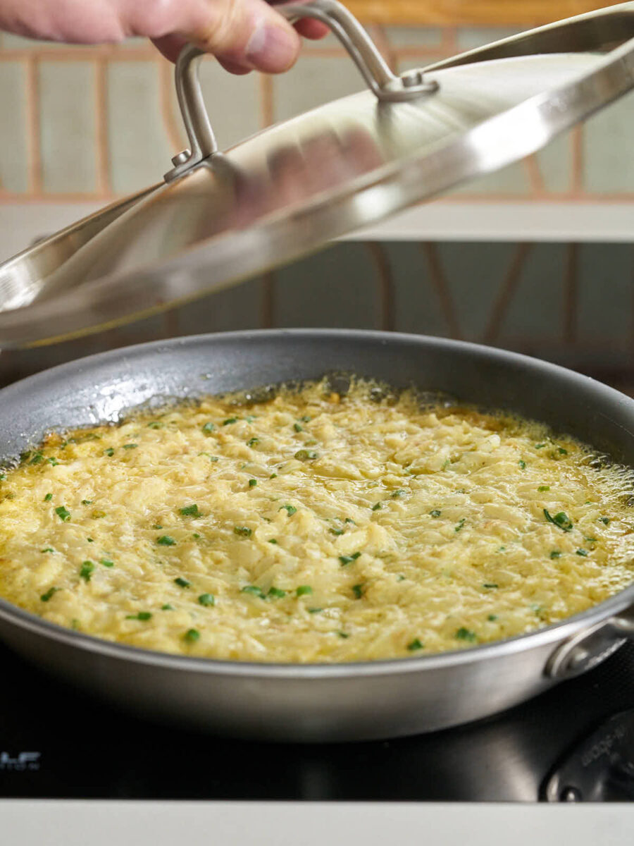 A person's hand lifting a stainless steel lid off a black frying pan, revealing a partially cooked frittata.