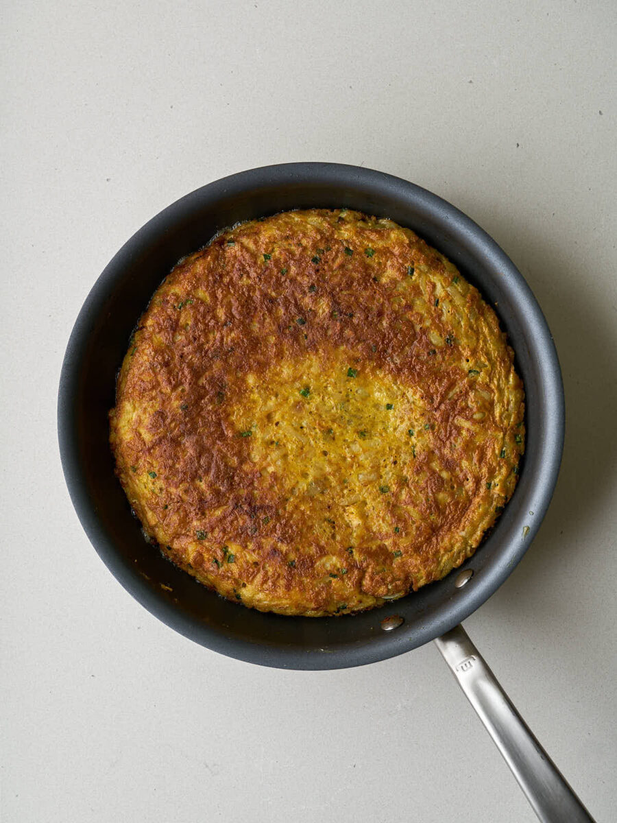 A golden brown, cooked kuku (potato frittata) in a black frying pan, viewed from directly above.