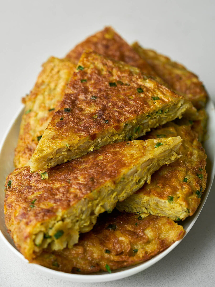 A close-up, angled view of an oval white platter piled with golden brown triangular slices of kuku (potato frittata).