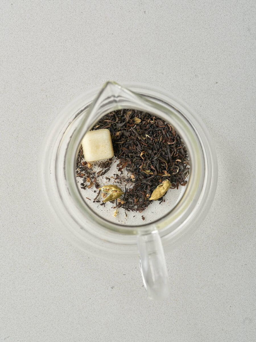 Overhead view of a clear glass teapot containing dry black tea leaves, two green cardamom pods, and a single white sugar cube.
