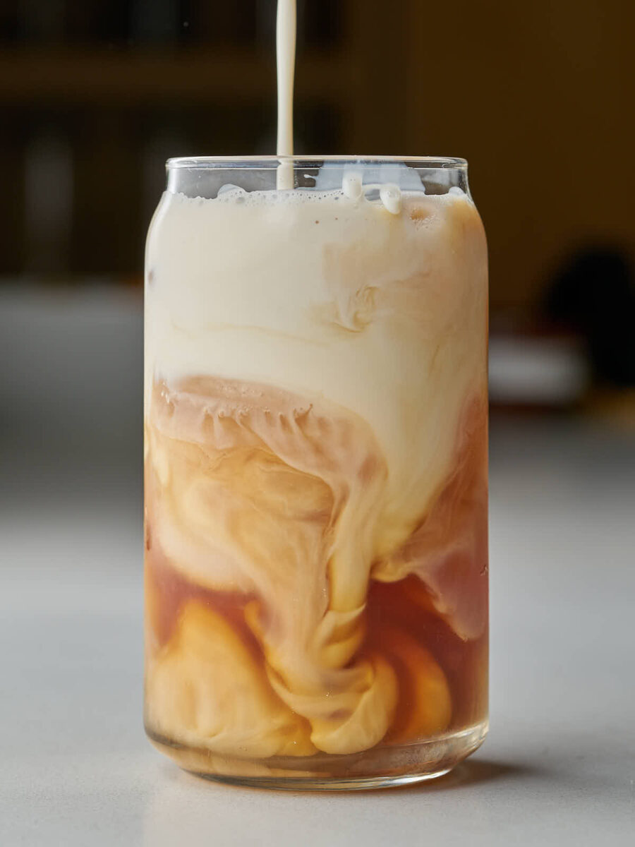 A clear glass filled with ice and iced tea, with a stream of white milk being poured into it, creating swirls of light brown and white as the liquids mix.