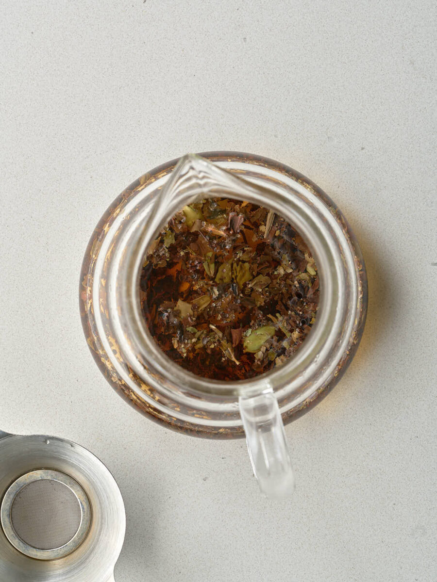 Overhead view of a clear glass teapot after hot water has been added, showing the steeped black tea leaves, cardamom pods, and dissolved sugar cube in the amber liquid. A metal tea strainer sits next to the teapot.