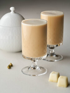 Two clear, footed glasses with etched floral patterns, filled with a light brown, creamy Persian latte. To the left is a white lidded sugar bowl, and to the right are two white sugar cubes.