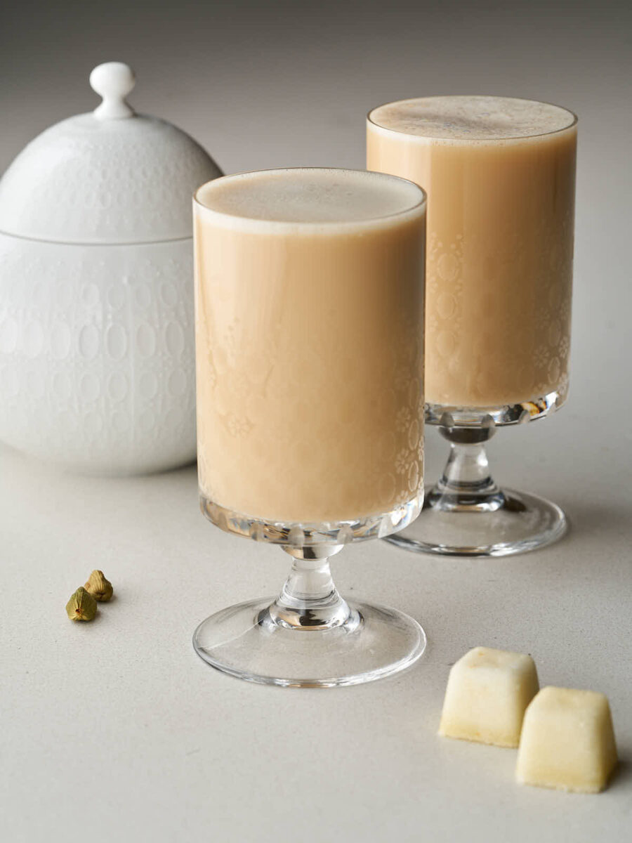 Two clear, footed glasses with etched floral patterns, filled with a light brown, creamy Persian latte. To the left is a white lidded sugar bowl, and to the right are two white sugar cubes.