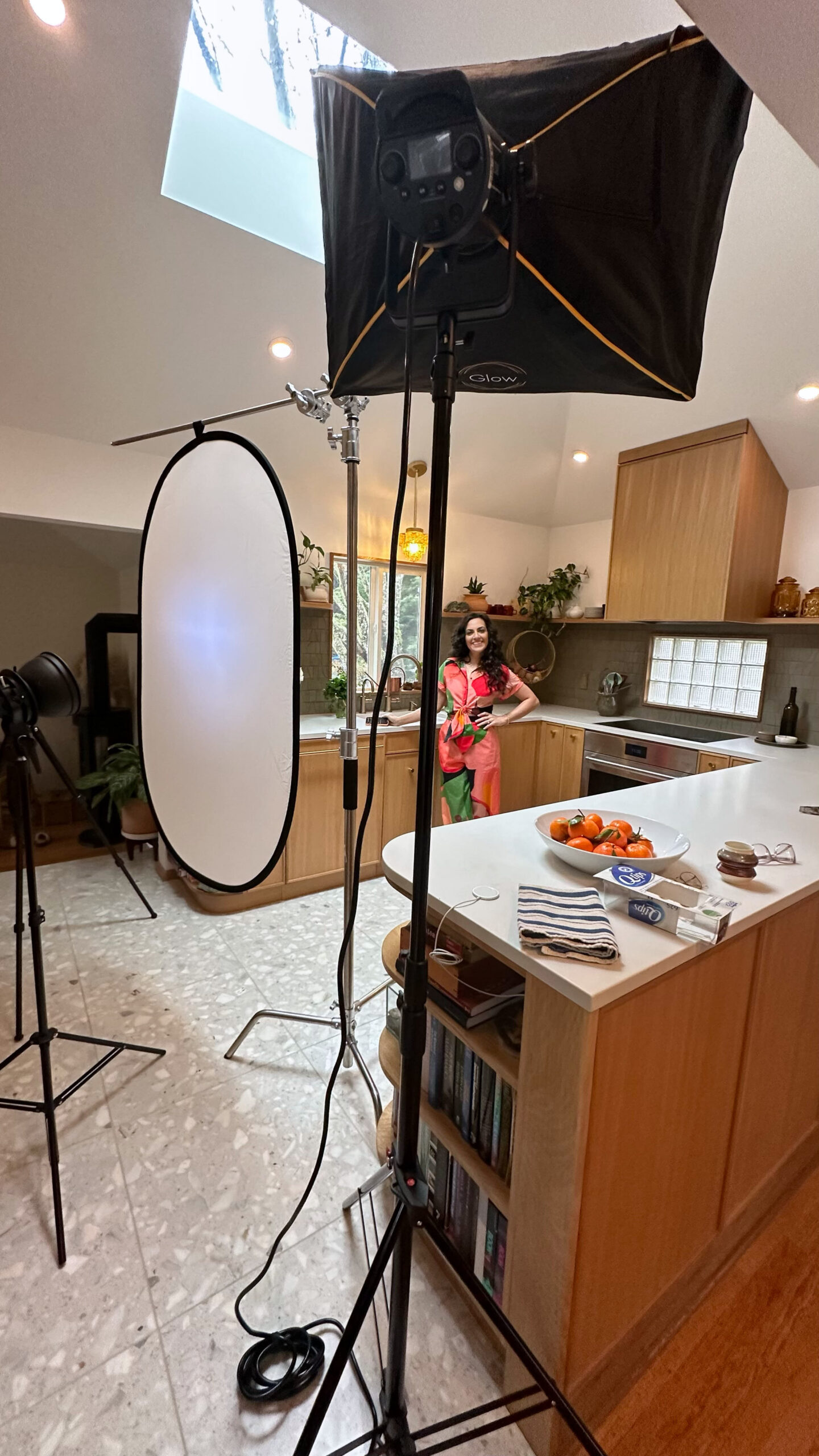 A kitchen photoshoot with professional lighting. A woman in colorful clothing stands in the background, while a large softbox and diffusion panel are in the foreground.