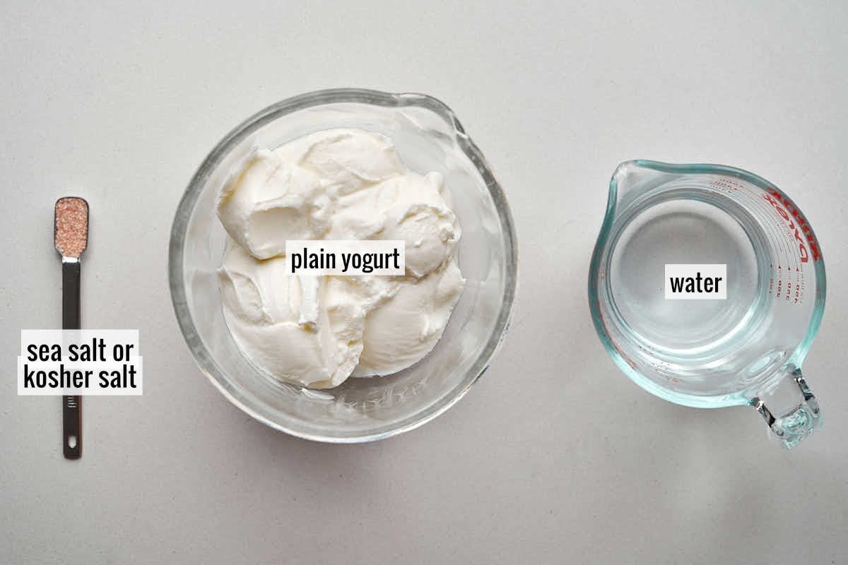 Three ingredients for a recipe are laid out on a light gray surface: a small metal spoon with pink sea salt or kosher salt, a clear glass bowl filled with plain yogurt, and a clear glass measuring cup with water.