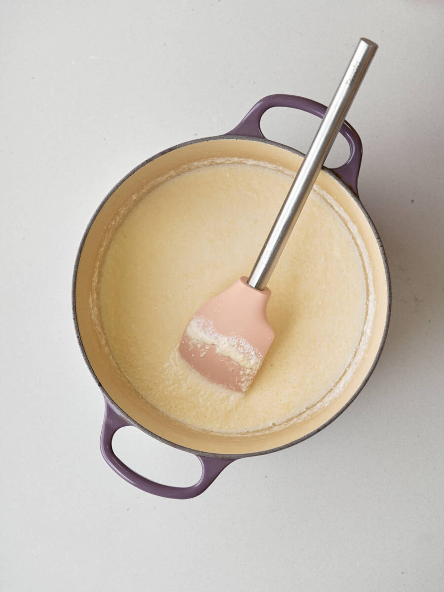 A pale yellow, slightly lumpy liquid fills a purple-handled pot. A pink silicone spatula with a metal handle rests in the liquid.