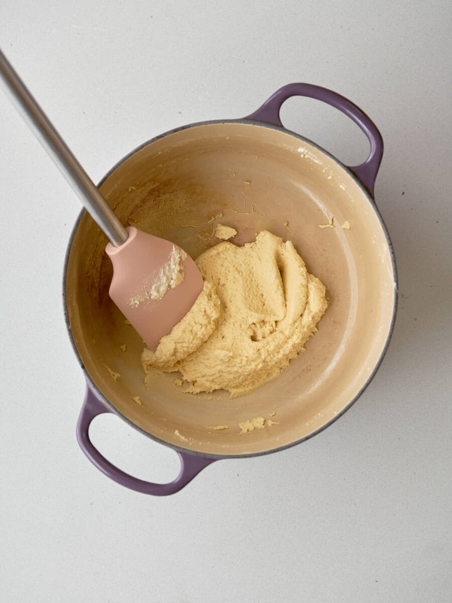 A light-colored, thick paste-like substance is being scraped from the side of a purple-handled pot with a pink silicone spatula.