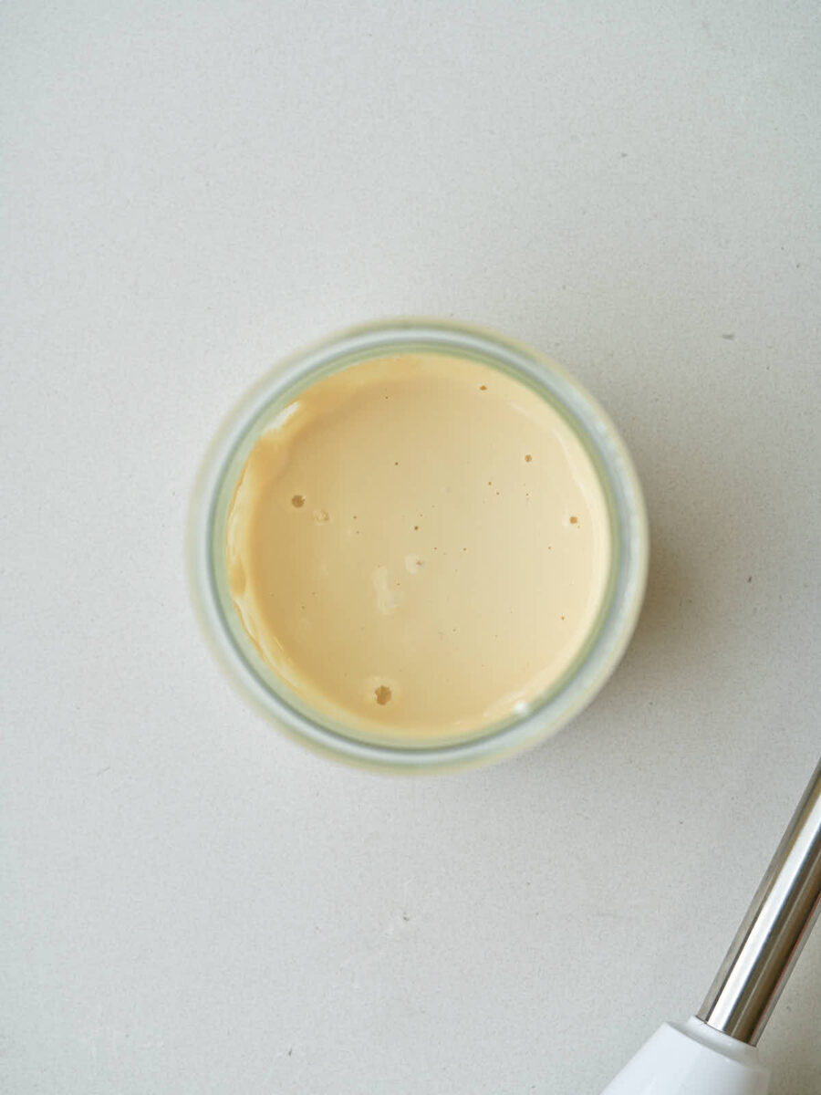 A top-down view of a clear glass jar filled with a smooth, pale tan liquid. The tip of a metal immersion blender stick is visible in the bottom right corner of the frame.