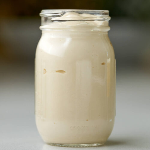 A clear glass jar filled with a thick, creamy, pale tan liquid. The jar sits on a light-colored surface with a blurred background.