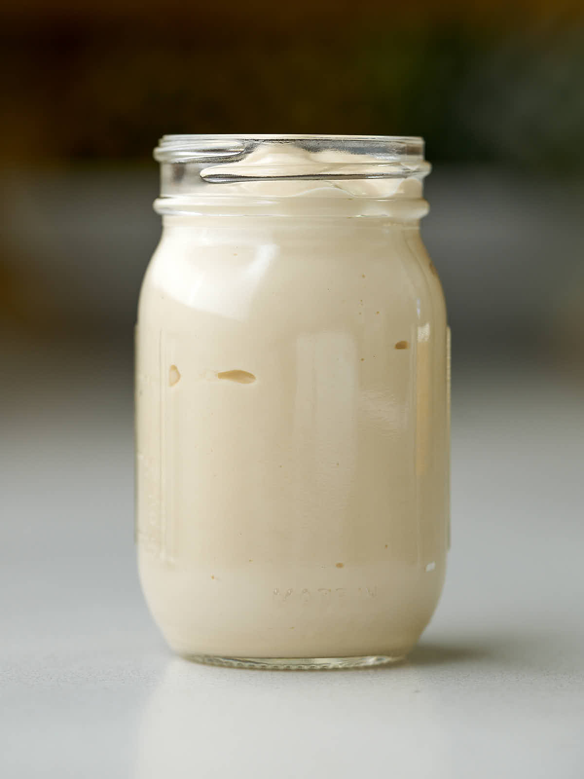 A clear glass jar filled with a thick, creamy, pale tan liquid. The jar sits on a light-colored surface with a blurred background.