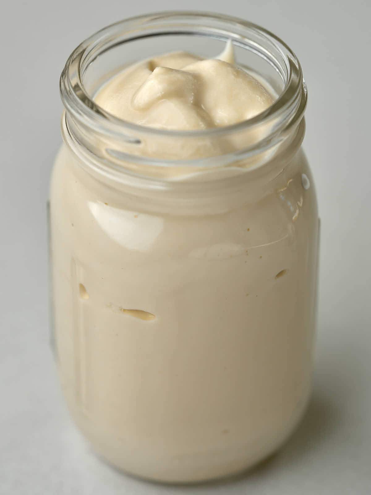 A clear glass jar is filled with a thick, creamy, pale tan substance, mounded slightly at the top. The jar sits on a light-colored surface with a blurred background.