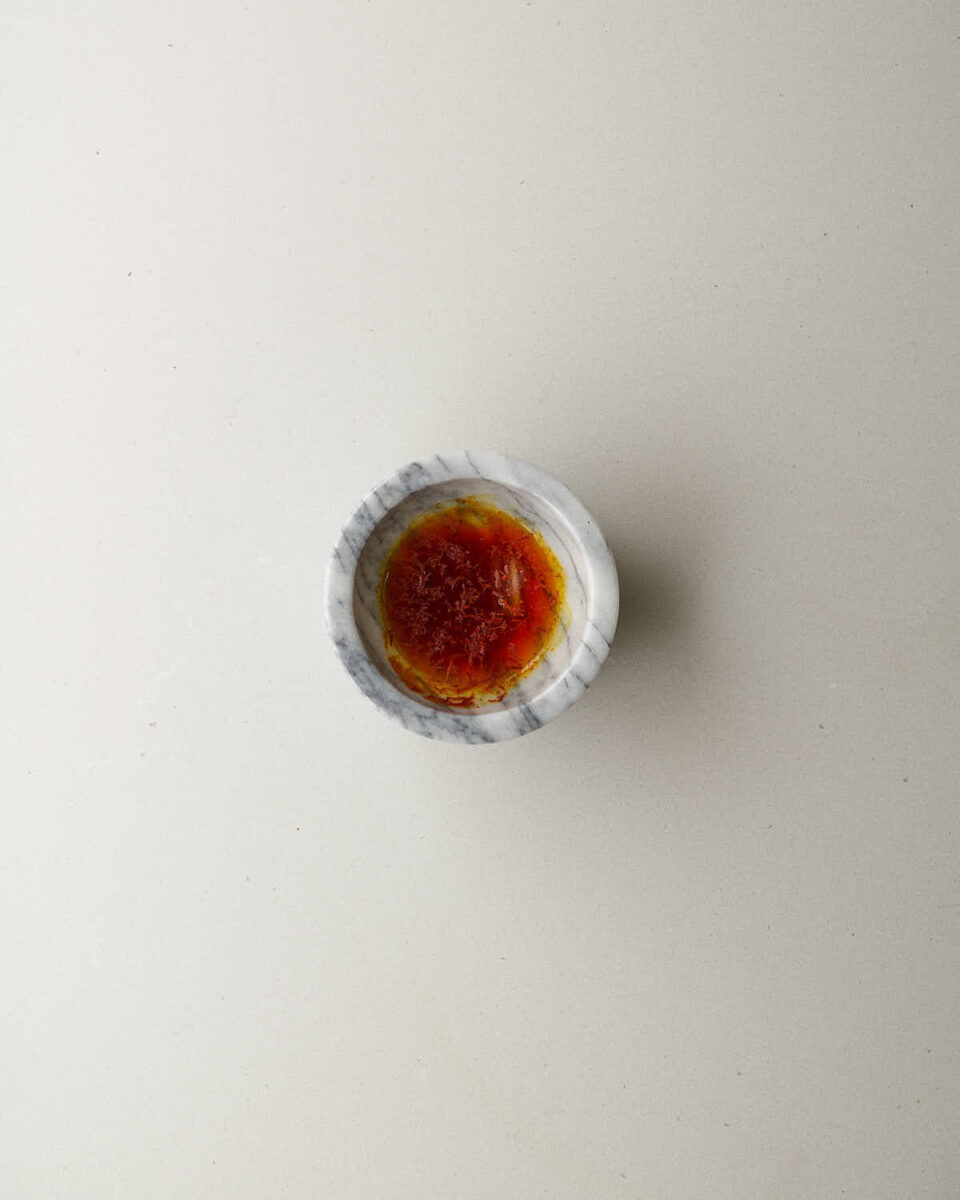 A close-up, overhead shot of a small marble bowl with saffron threads soaking in a reddish-orange liquid.
