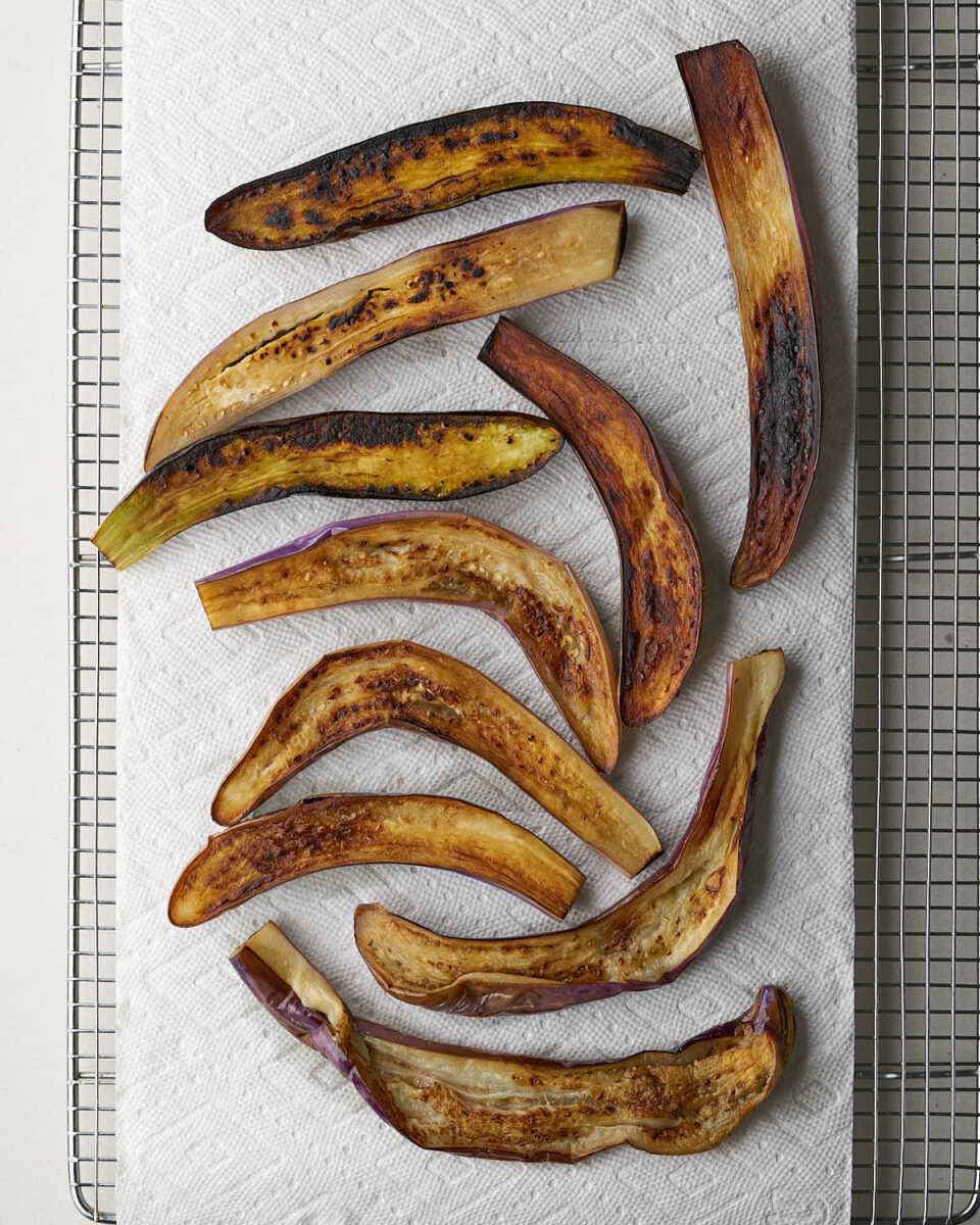 Overhead shot of roasted eggplant slices resting on a paper towel-lined wire rack. The eggplant skins are dark purple and some are charred.