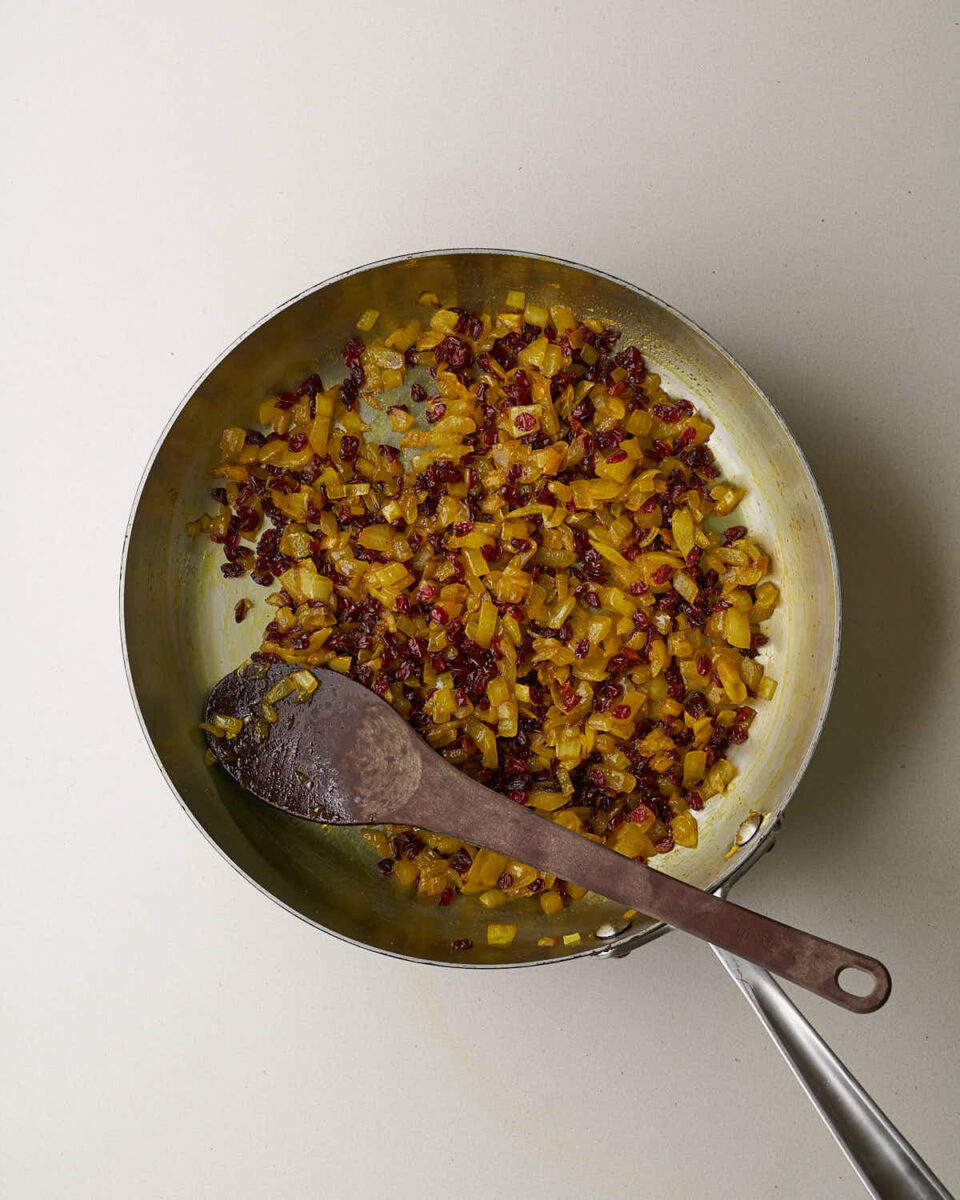 An overhead shot of a skillet with a mix of cooked, diced yellow onions and red barberries. A wooden spoon rests in the skillet.