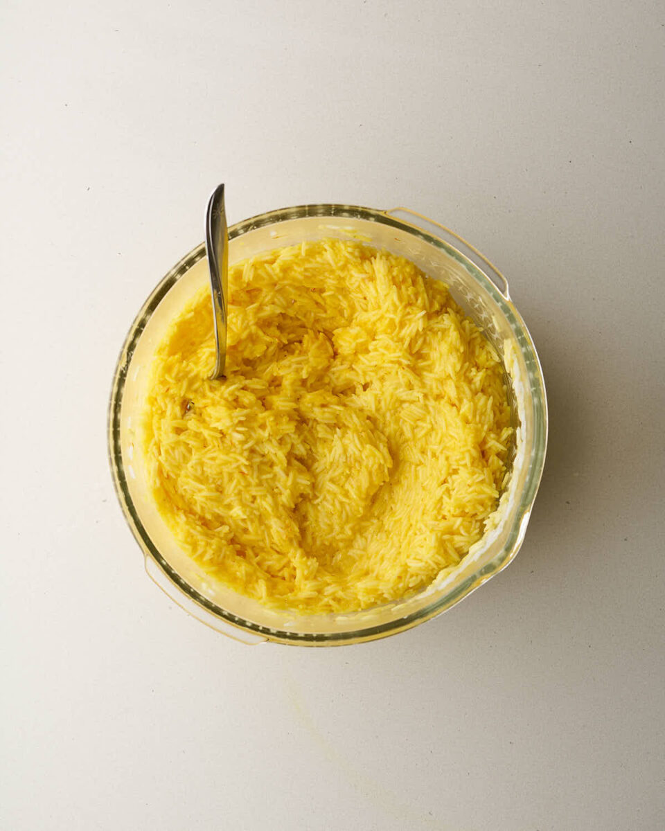 An overhead shot of a glass bowl filled with cooked rice that has been mixed with a yellow sauce, making the rice look light yellow. A metal spoon is in the bowl.