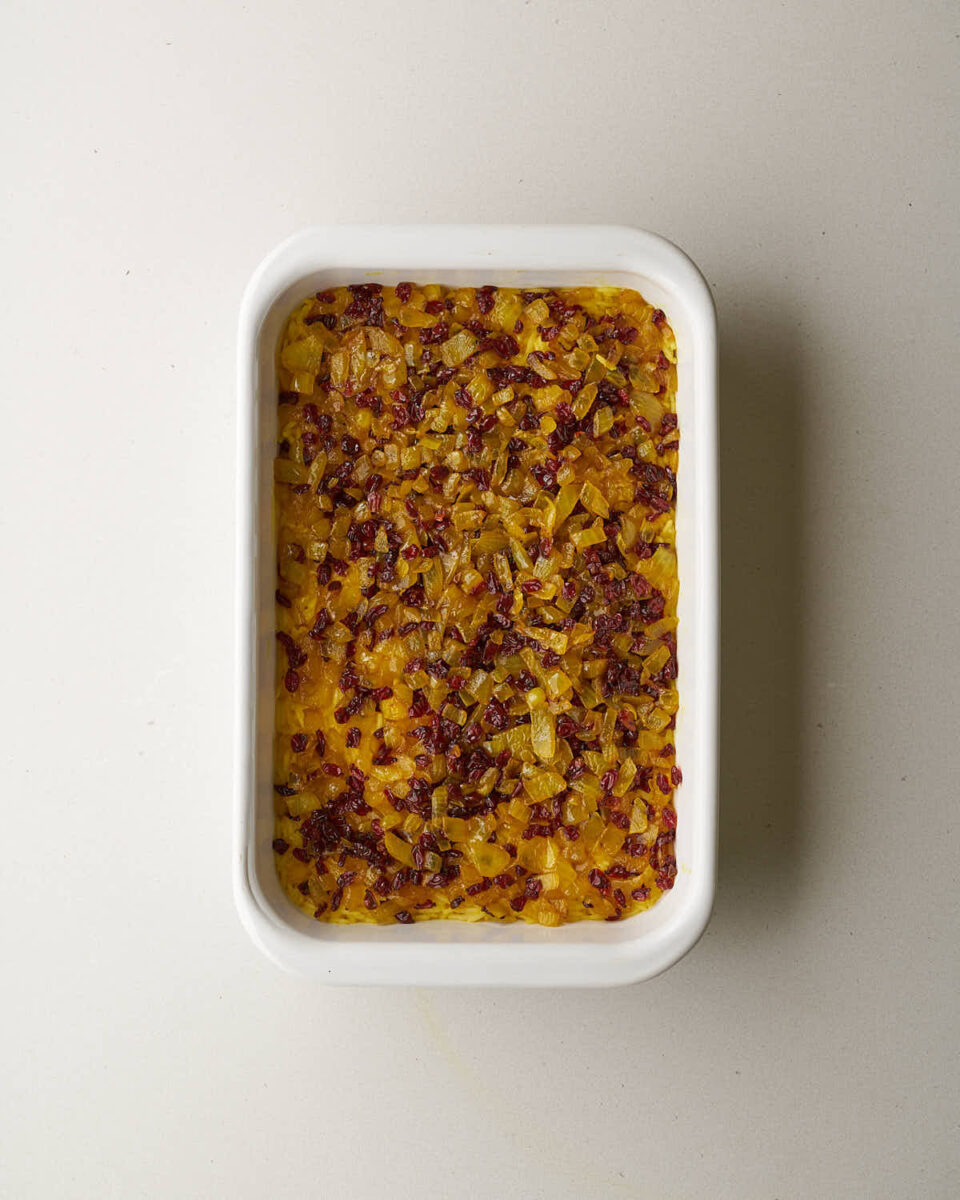 An overhead shot of a white rectangular baking dish. A layer of yellow rice is topped with a layer of cooked onions and barberries.
