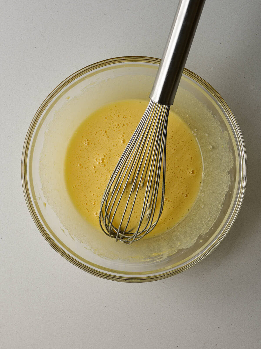A glass bowl contains four egg yolks and a milky liquid mixture. A silver whisk is resting inside the bowl.