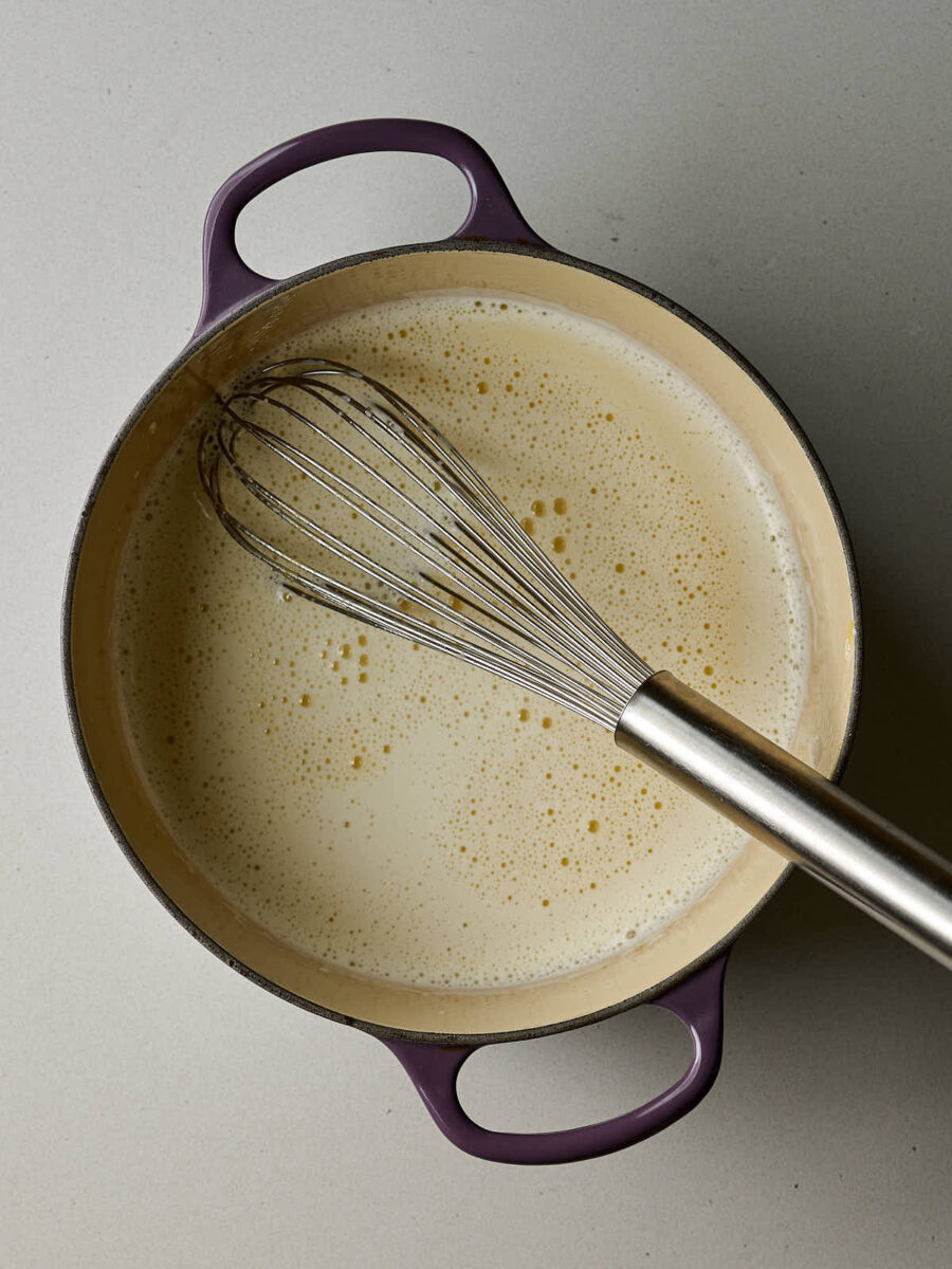 A purple cast iron pot contains a pale yellow liquid. A silver whisk is resting in the liquid.