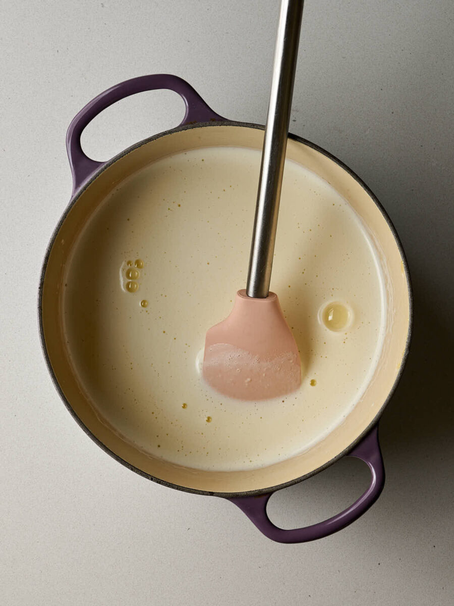 A purple cast iron pot contains a pale yellow liquid. A pink spatula with a silver handle is resting in the liquid.