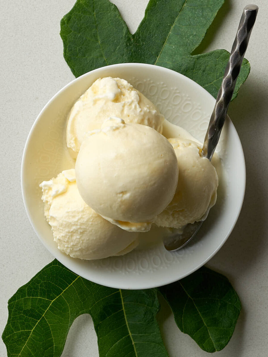 An overhead shot of a white bowl filled with four scoops of pale yellow ice cream. A fig leaf is placed under the bowl to the left and another to the right. A silver spoon with a textured handle rests in the bowl on the right side.