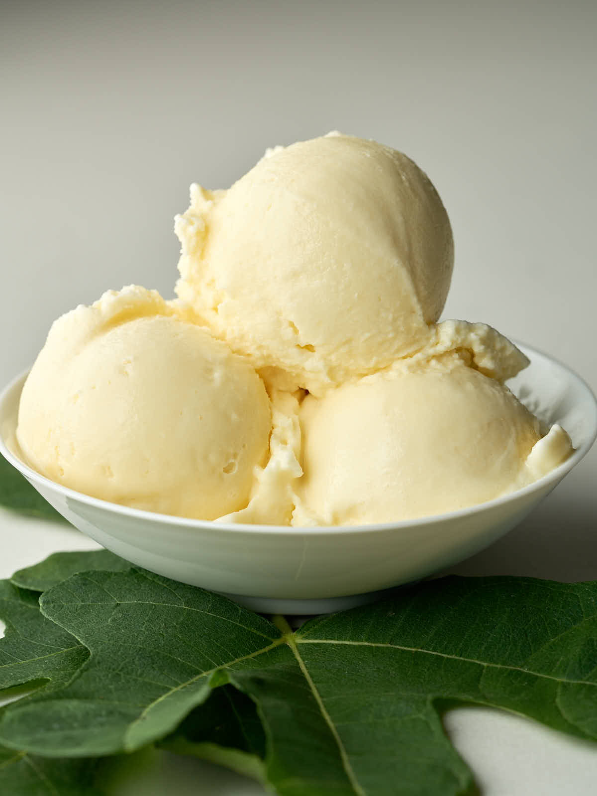 Three scoops of pale yellow ice cream sit in a white bowl, with a few large green fig leaves arranged beneath it on a gray surface.