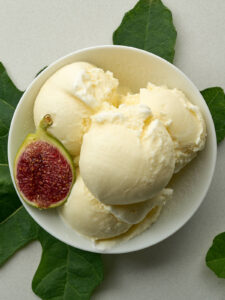 An overhead shot of a white bowl filled with four scoops of pale yellow ice cream and half a fig. A fig leaf is placed under the bowl to the left and another to the right.