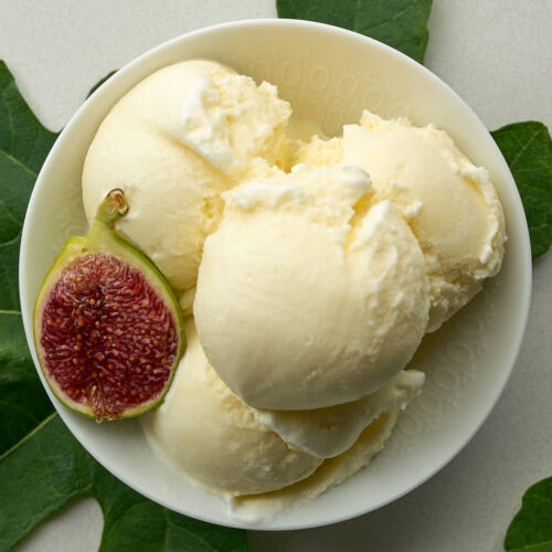 An overhead shot of a white bowl filled with four scoops of pale yellow ice cream and half a fig. A fig leaf is placed under the bowl to the left and another to the right.