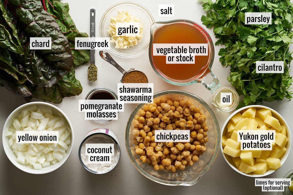 Overhead shot of ingredients for Shawarma Chickpea Stew laid out, including chopped onions, chard, dried spices, pomegranate momlasses, coconut cream in a can, vegetable broth, oil, chickpeas, chopped garlic, chopped potatoes, and fresh herbs.
