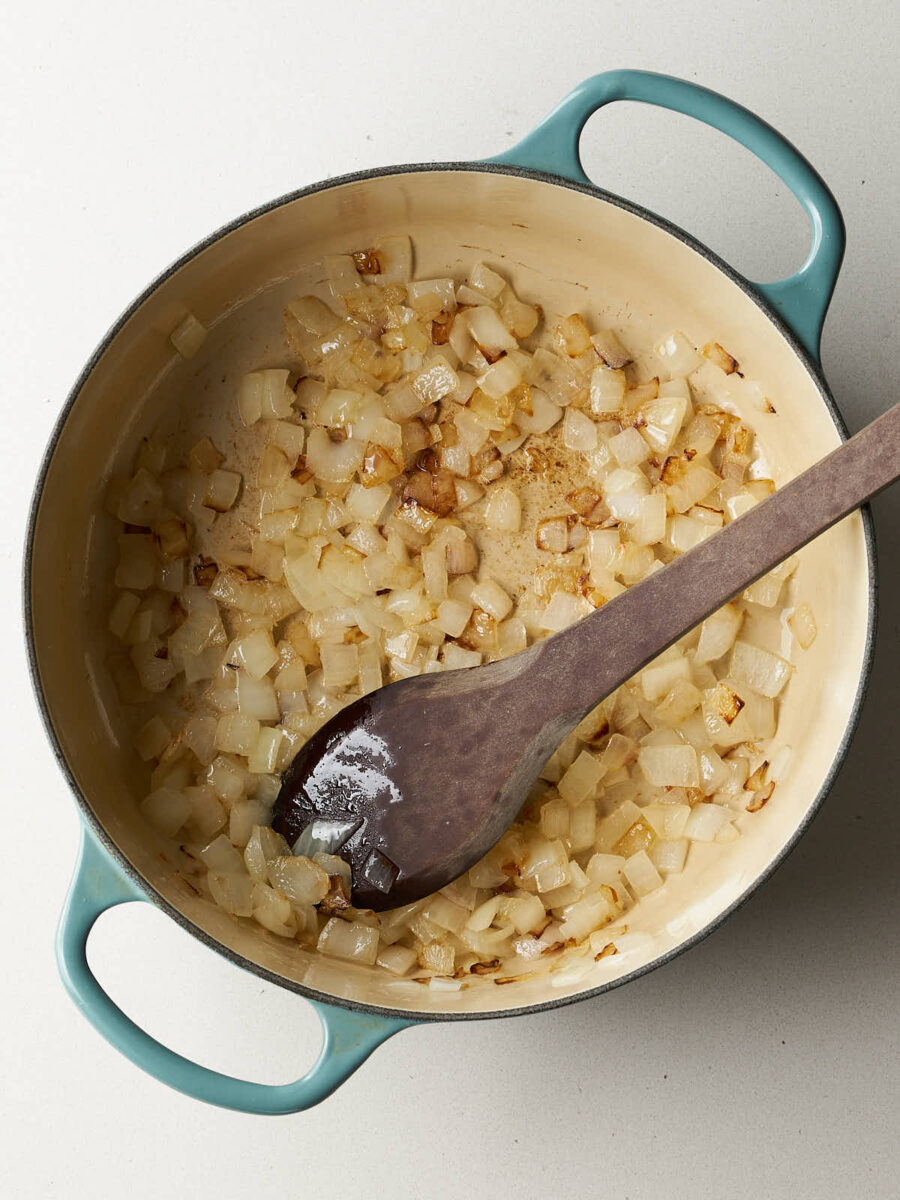 Close-up of diced onions being sautéed in a light blue dutch oven with a wooden spoon.