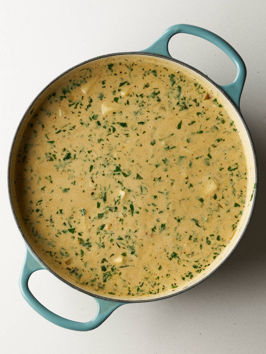 A creamy, yellowish stew base with small chunks of potatoes, chickpeas, and flecks of green herbs in a light blue dutch oven.